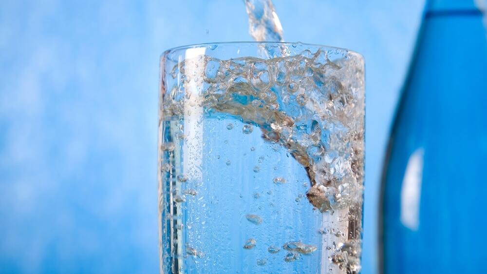 Glas vor blauem Hintergrund wird mit frischem, sprudelndem Trinkwasser befüllt.