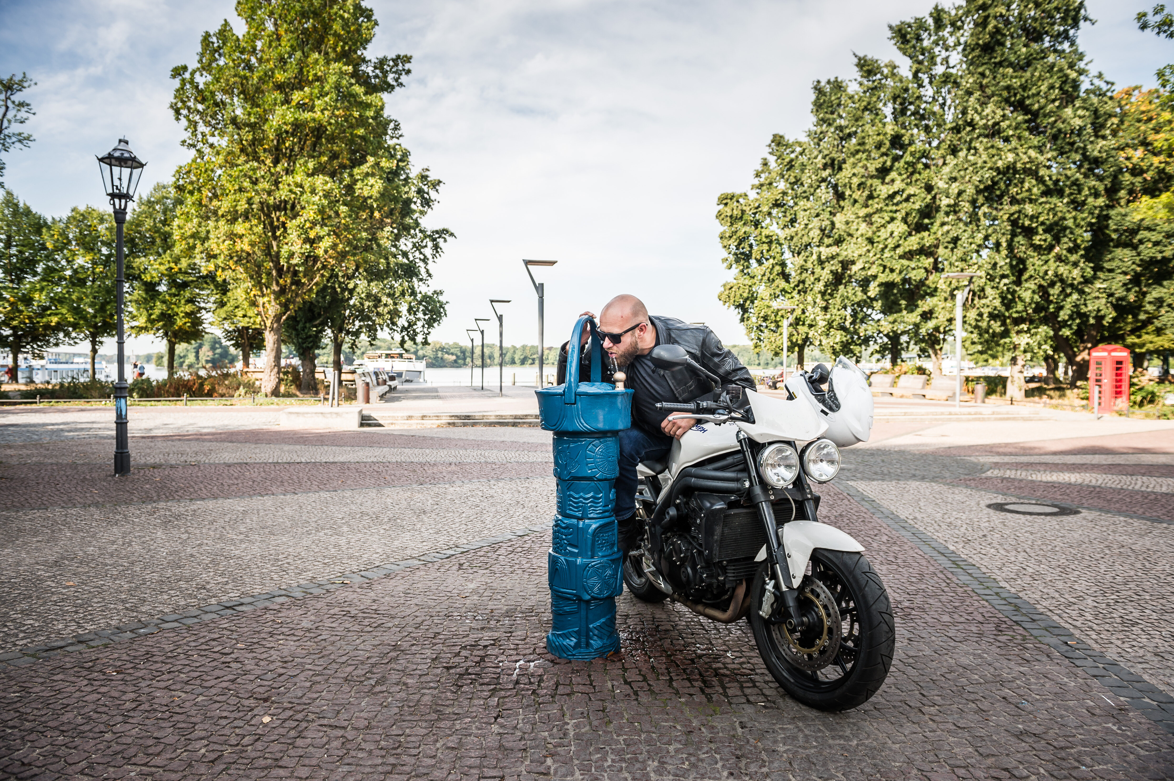 Motorradfahrer hält an einem öffentlichen Trinkwasserbrunnen und trinkt dort aus dem Hahn.